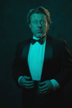 Smokin ve papyon takan sarışın adam. Mavi ve turuncu ışıkla karanlık duvara karşı.