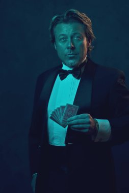 Smokinli ve papyonlu bir adam poker kartı tutuyor. Koyu bir duvarın önünde mavi ve turuncu ışıkla aydınlandı..