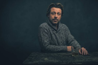 Eski ahşap masanın arkasında bir bardak viskiyle gri yün kazaklı bir adam..