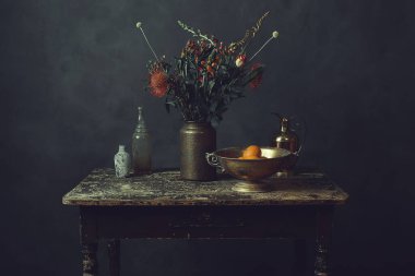 Üzerinde çiçekler olan eski ahşap bir masa, portakallı bronz bir kase, bir sürahi ve iki cam şişe..