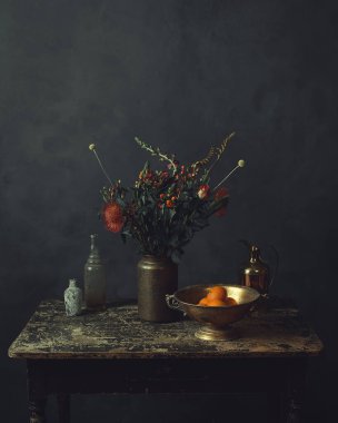 Üzerinde çiçekler olan eski ahşap bir masa, portakallı bronz bir kase, bir sürahi ve iki cam şişe..