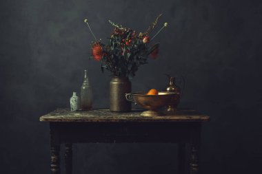 Üzerinde çiçekler olan eski ahşap bir masa, portakallı bronz bir kase, bir sürahi ve iki cam şişe..