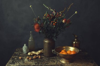 Üzerinde çiçekler, zencefil ve ceviz olan eski ahşap bir masa, portakallı bronz bir kase, bir sürahi ve iki cam şişe..