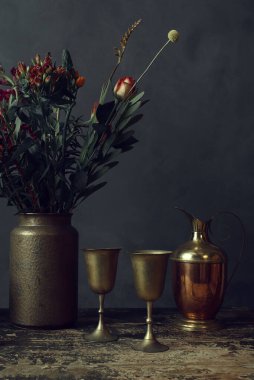 Çiçekli çömlek vazosuyla kaplı eski ahşap bir masa, iki bronz bardak ve bir sürahi..