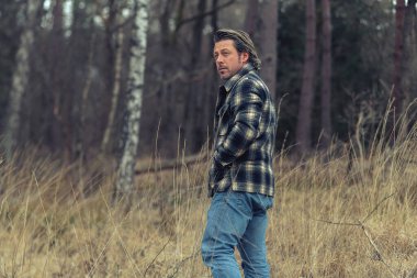 Tarlada kareli ceketli bir adam. Ormanın yakınında uzun sarı çimenler var..