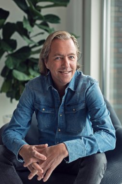 Cheerful middle aged blonde man in jeans shirt sits in chair near window of living room.