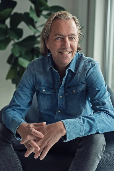 Cheerful middle aged blonde man in jeans shirt sits in chair near window of living room.