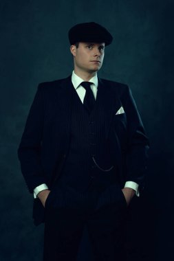 Koyu renk çizgili takım elbiseli ve koyu renkli duvarın önünde şapka takan adam..