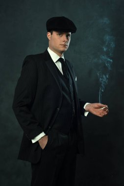Koyu renk çizgili takım elbiseli, elinde bir sigarayla siyah bir duvarın önünde duran klasik bir adam..