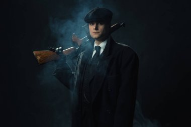 Siyah takım elbiseli, şapkalı, dumanlı, siyah bir duvarın önünde duran eski tip bir adam omzunda bir tüfek tutuyor..