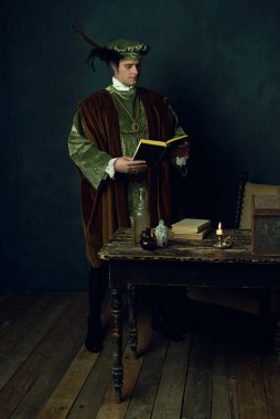 Elinde kitap olan bir Rönesans adamı üzerinde kitaplar, üç cam şişe ve bir şamdan olan bir masanın arkasında duruyor..