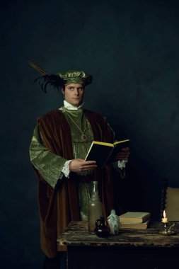 Elinde kitap olan bir Rönesans adamı üzerinde kitaplar, üç cam şişe ve bir şamdan olan bir masanın arkasında duruyor..