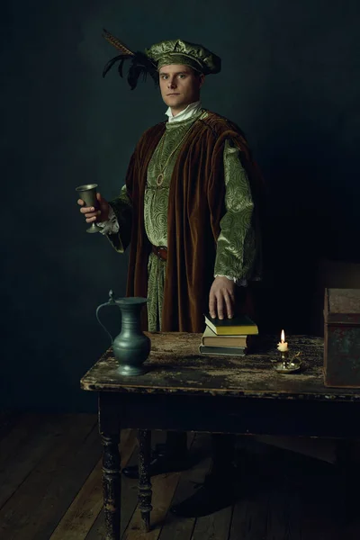 Renaissance man with a tin goblet stands behind a table with books on it, a tin pitcher, a candlestick and an iron box.