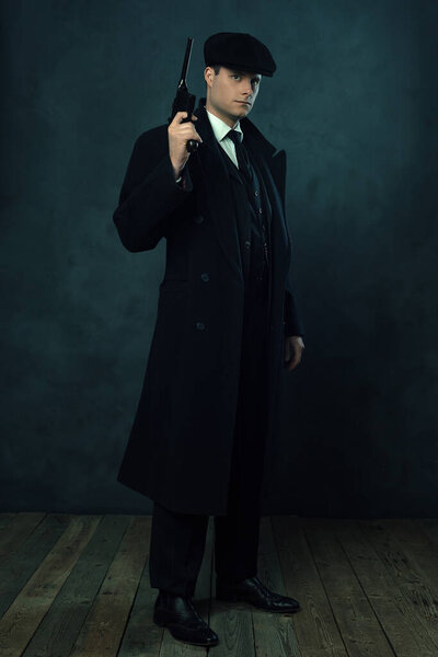 Menacing vintage man in a dark striped suit and a cap holding a handgun in front of a dark wall. 