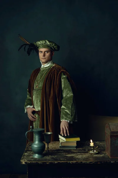 Renaissance man stands behind a table with books on it, a tin pitcher, a tin goblet, a candlestick and an iron box.