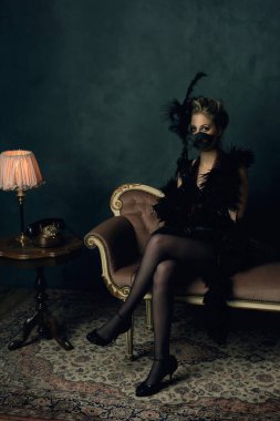 Siyah boa 'lı, retro-yatak elbiseli, kolyeli, antika bir koltukta, masa lambası ve telefonu olan bir sehpanın yanında oturan baştan çıkarıcı genç kadın..