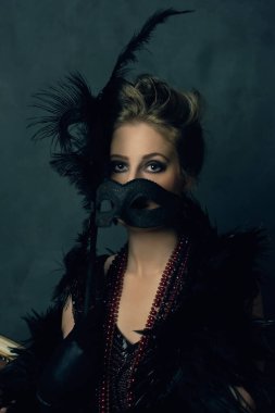 Esrarengiz baştan çıkarıcı kadın siyah bir boa ve antika bir kanepede oturan kolyeli retro bir maske takıyor..