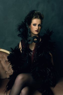 Esrarengiz baştan çıkarıcı kadın siyah bir boa ve antika bir kanepede oturan kolyeli retro bir maske takıyor..