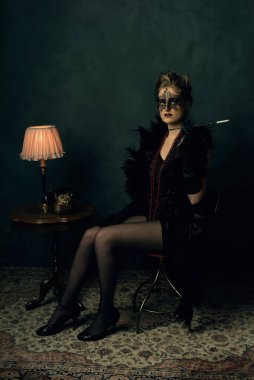 Siyah boa ve maskeli esrarengiz bir genç kadın. Masa lambası ve telefonu olan kahve masasının yanında kolyeli..