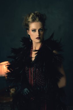 Eskimiş erkek elbiseli, kolyeli baştan çıkarıcı genç bir kadın masa lambası ve telefonlu bir sehpanın önünde duruyor..