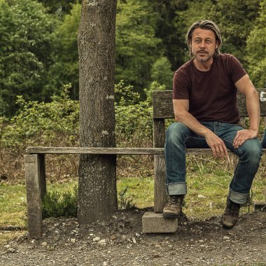 Kot pantolonlu, kahverengi tişörtlü, sarı saçlı bir adam ahşap bir bankta oturuyor..