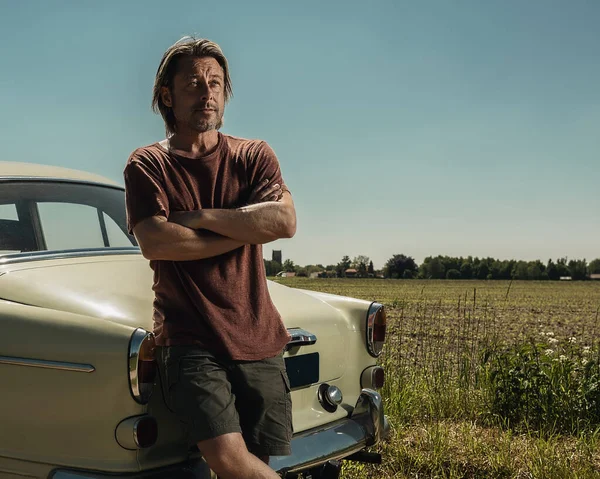 Blond man in t-shirt and shorts standing by the trunk of a classic car in sunny countryside.