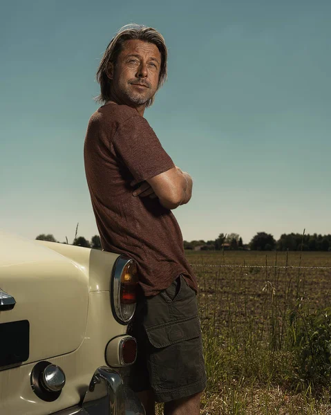 Tシャツを着たブロンドの男と日当たりの良い田舎の古典的な車のトランクのそばに立つショートパンツ — ストック写真
