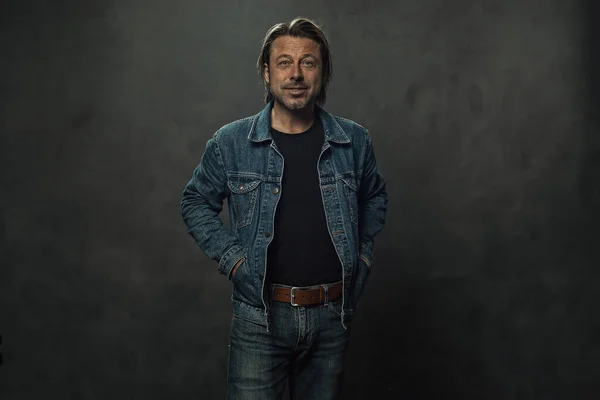 ダークグレーの壁の前に茶色の革ベルトとデニムジャケットを着たジーンズのブロンドの男 — ストック写真