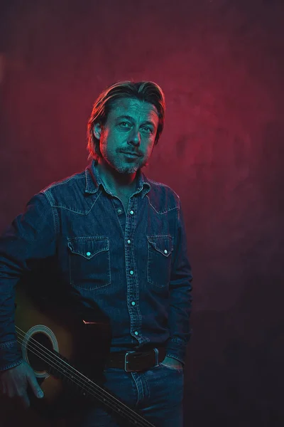 Man with acoustic western guitar in jeans shirt and jeans with belt in red and blue colored light.