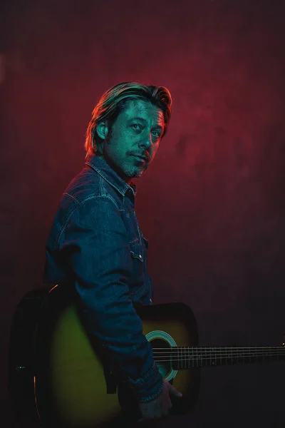 Man with acoustic western guitar in jeans shirt and jeans with belt in red and blue colored light.