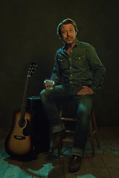 音乐家坐在木凳上 手里拿着威士忌 旁边拿着一把吉他和一个吉他盒 — 图库照片