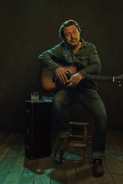 Musician sitting on a wooden stool with an acoustic guitar and a cigarette with next to him a guitar case with a glass of whiskey and an ashtray.