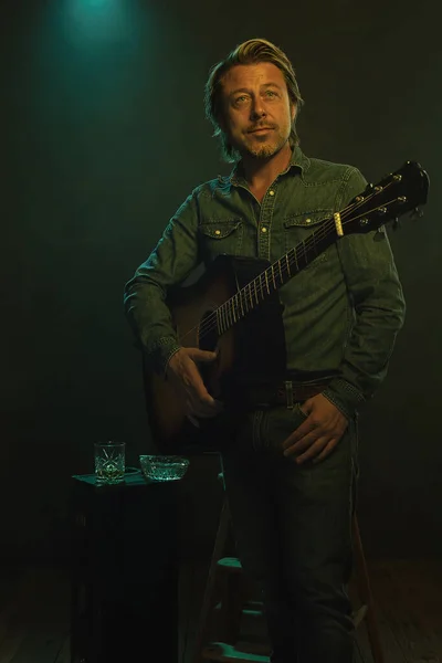 Country and western musician in jeans and denim shirt stands with an acoustic guitar in front of a wooden stool and a guitar case with a glass of whiskey and an ashtray on top.