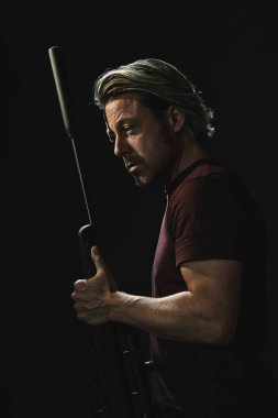 Koyu kahverengi tişörtlü, elinde tüfekle omzunun üstünden bakan sarı saçlı adamın koyu renkli portresi..