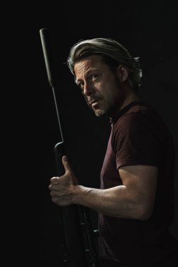 Koyu kahverengi tişörtlü, elinde tüfekle omzunun üstünden bakan sarı saçlı adamın koyu renkli portresi..