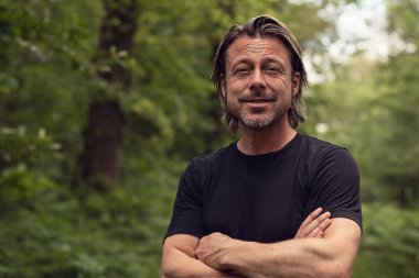 Çapraz kollu siyah tişörtlü, gür ormanda gülümseyen sarışın adam..