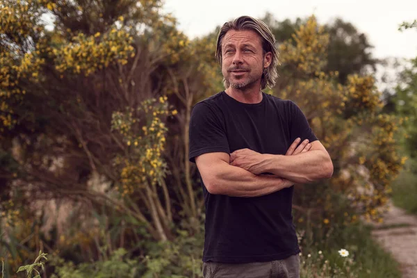 Smiling blond man with crossed arms and stubble beard in a black t-shirt stands in front of a bush with yellow flowers.