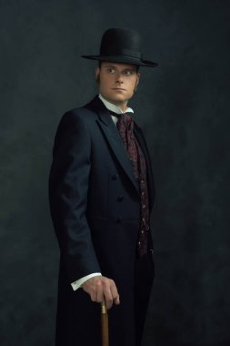 Viktorya tarzı şık giyinmiş genç bir adam. Koyu gri bir duvarın önünde bastonla dikiliyorum..