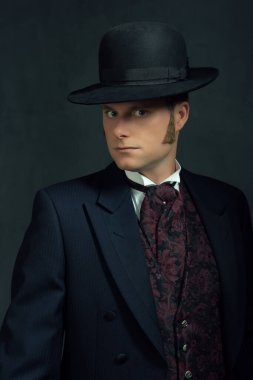 Viktorya tarzı şık giyinmiş genç bir adam. Koyu gri bir duvarın önünde duruyor..