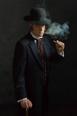 Puro tüttüren şık Viktorya dönemi kıyafetleriyle koyu gri bir duvarın önündeki bir odada duran genç adam..