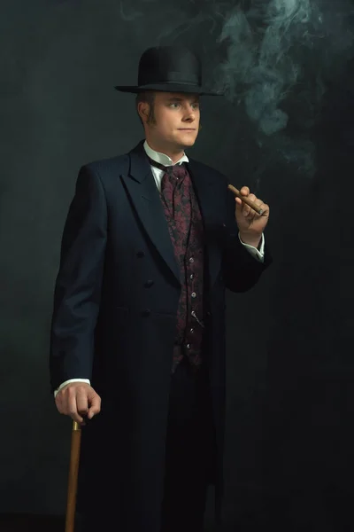 身穿维多利亚时代老式服装 头戴手杖 抽烟的雪茄青年男子站在深灰色墙壁前的房间里 — 图库照片