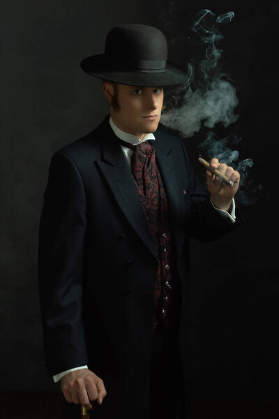 Cigar smoking young man in stylish vintage Victorian clothes with a walking stick stands in a room in front of a dark gray wall.