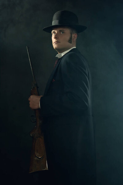 Shadowy portrait of a young man in vintage Victorian attire holding a rifle in a smoky room in front of a dark gray wall.