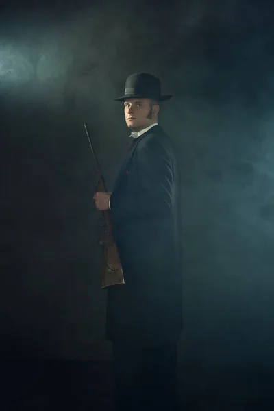 暗い灰色の壁の前のスモーキーな部屋にライフルを保持ヴィクトリアンの服装で若い男の影の肖像画 — ストック写真