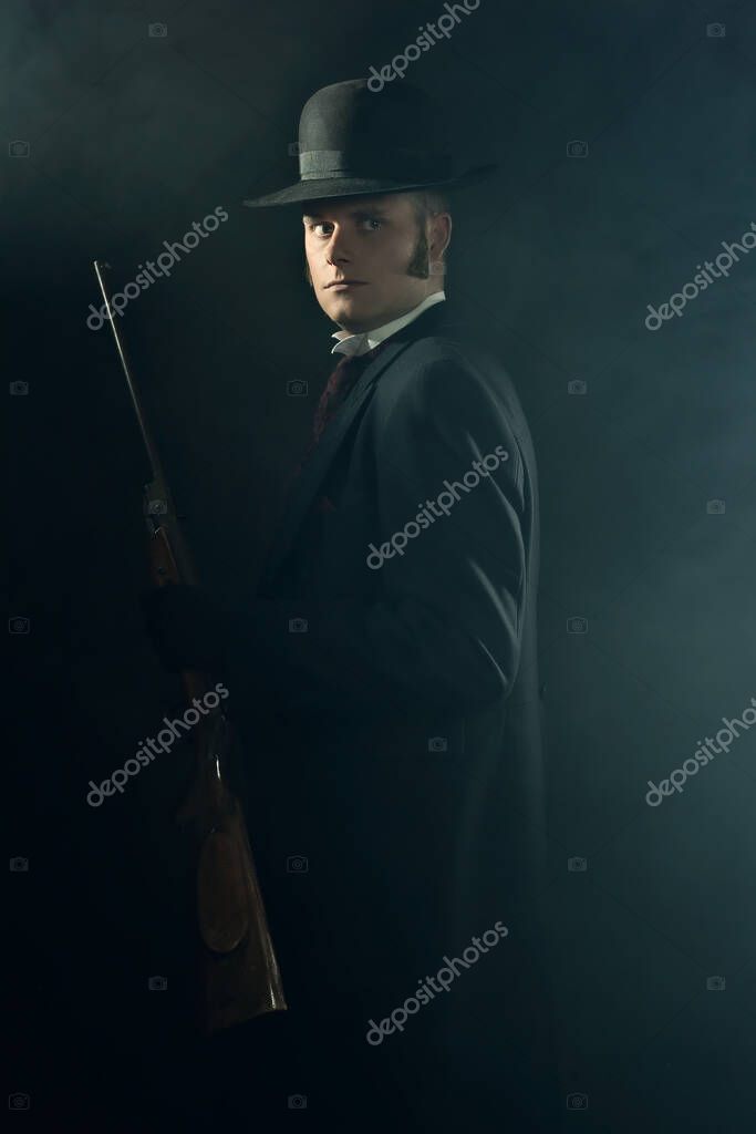 Shadowy portrait of a young man in vintage Victorian attire holding a rifle in a smoky room in front of a dark gray wall.