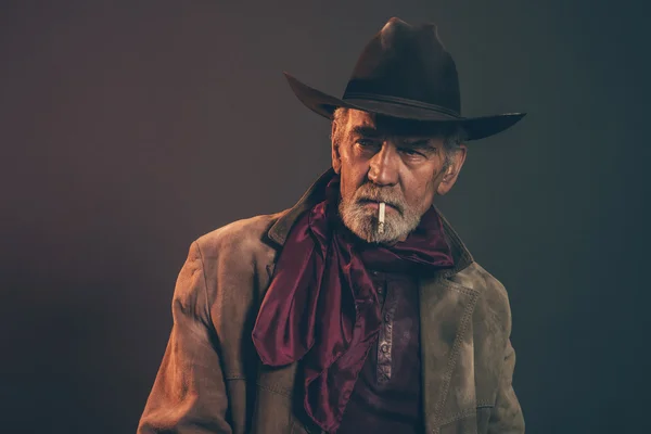 Old rough western cowboy with gray beard and brown hat smoking a — Stock Photo, Image