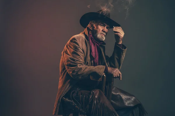 Viejo vaquero occidental áspero con barba gris y sombrero marrón fumar un —  Fotos de Stock