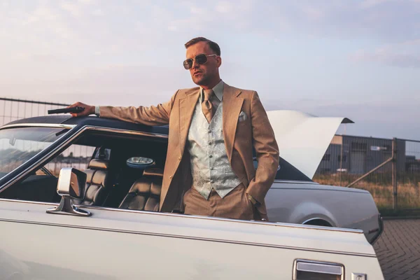 Retro 1970s gangster with pistol leaning against vintage car. — Stock Photo, Image