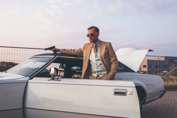 Retro 1970s gangster with pistol leaning against vintage car. — Stock Photo, Image