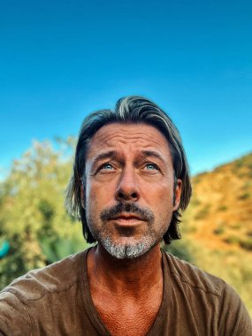 Middle aged man with blond half long hair and greyish beard outdoor in nature on a sunny day. Satisfied and relaxed expression on his face.